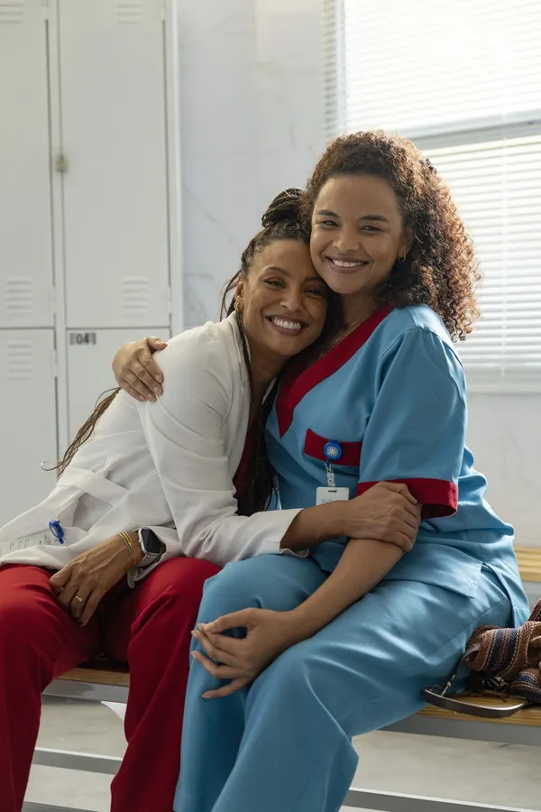 Bela, de jaleco branco, abraça Juli, de uniforme azul de enfermagem, num vestiário de hospital.