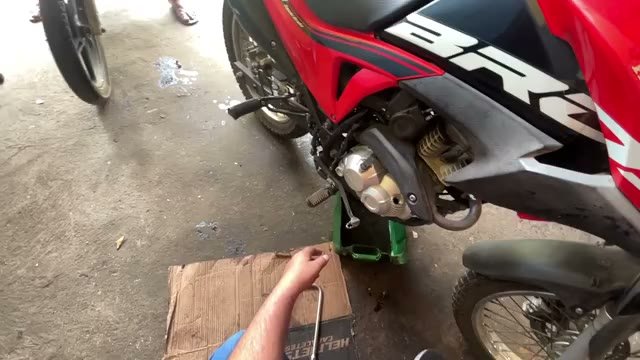 Oficina de moto vista em primeira pessoa: moto vermelha no cavalete, ferramentas sobre papelão no chão