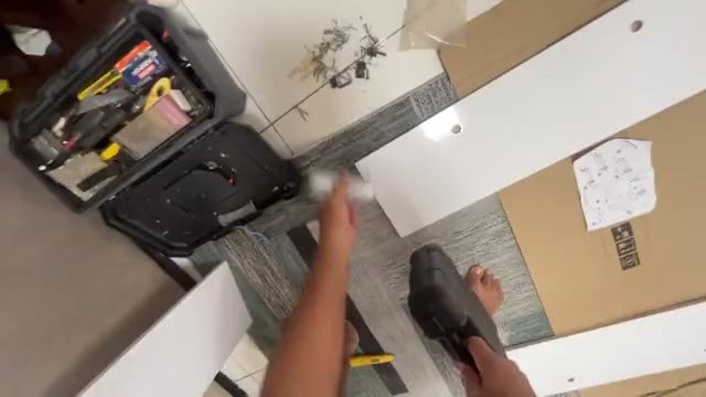 Montagem de móvel vista em primeira pessoa: caixa de papelão aberta, painéis brancos, manual e maleta de ferramentas no chão